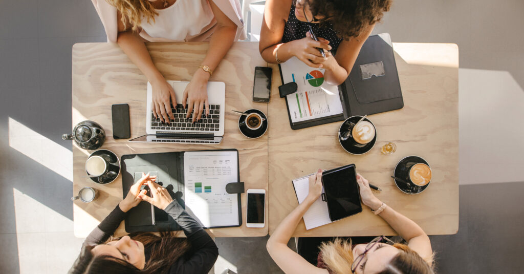 Top view of four women sitting around table in cafe with laptop, digital tablet and documents.