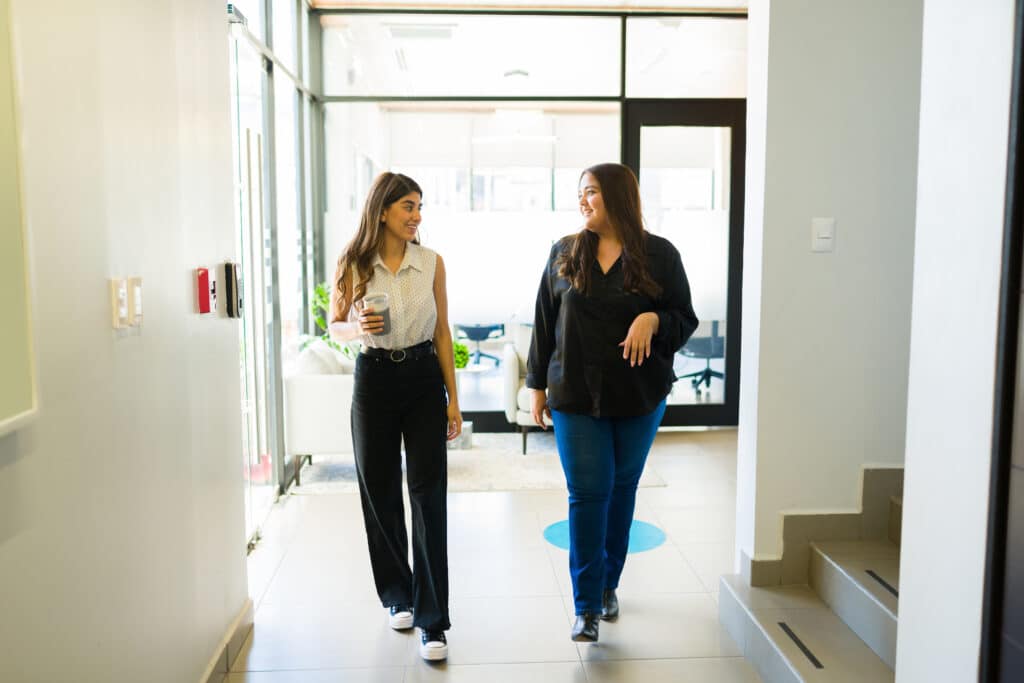 two women in business attire walk down a hallway while having a discussion