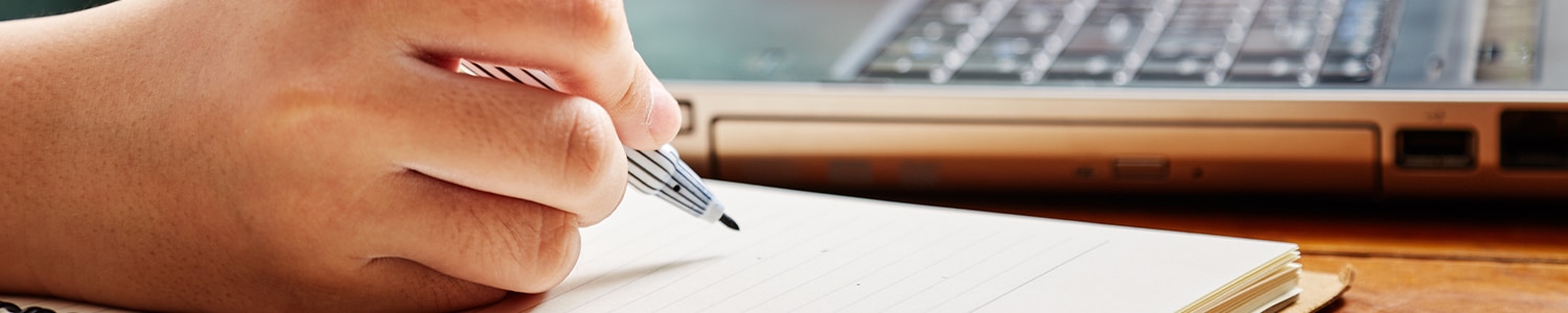 person writing in notebook next to laptop