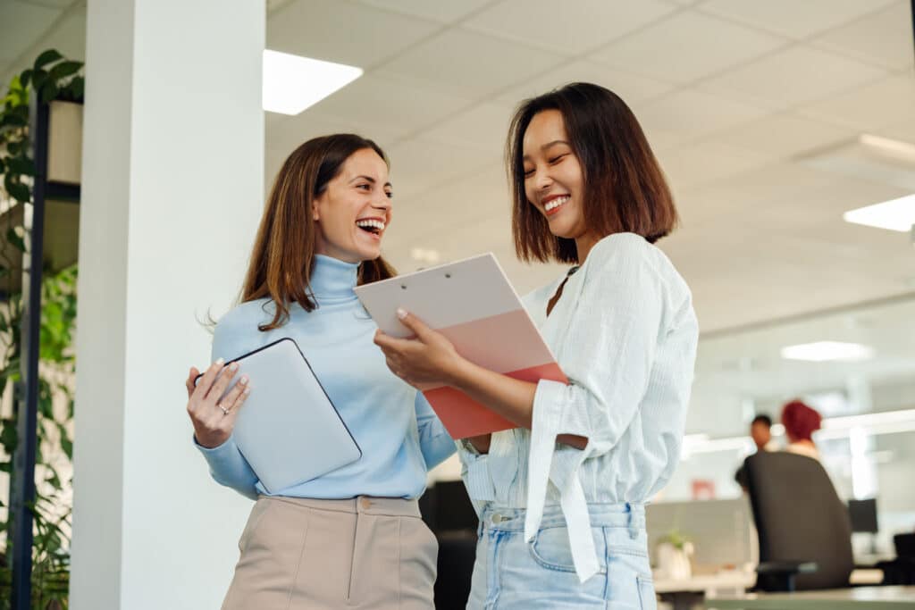 Two business woman chatting in an office while using tablet and a clipboard.