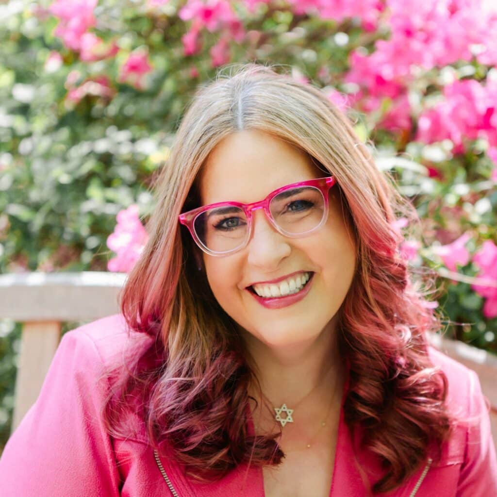 Headshot of Ahava Leibtage, CEO of Aha Media Group, wearing a pink jacket and with pink hair and pink glasses.